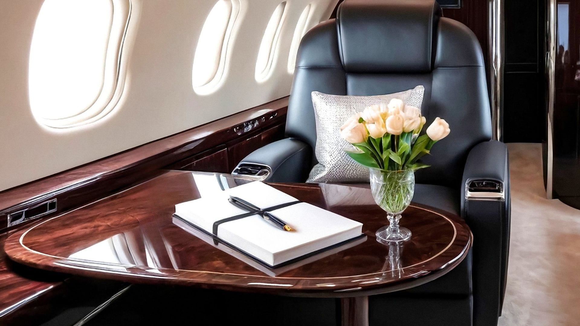Interior de cabina de jet privado con asiento en piel negra, mesa de madera pulida, libro de visitas blanco y jarrón de cristal con tulipanes blancos.